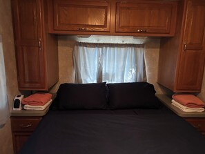 A compact bedroom with a bed, wooden cabinets, a window with curtains, and a bedside table with a phone and towels. - RV Retreat near Chemung River (Elmira)