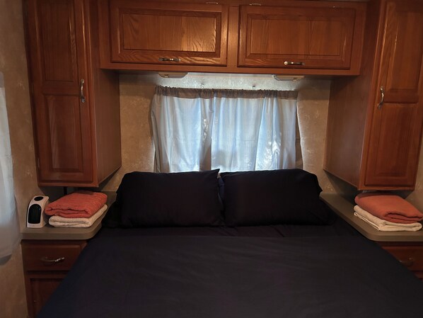 A compact bedroom with a bed, wooden cabinets, a window with curtains, and a bedside table with a phone and towels.
