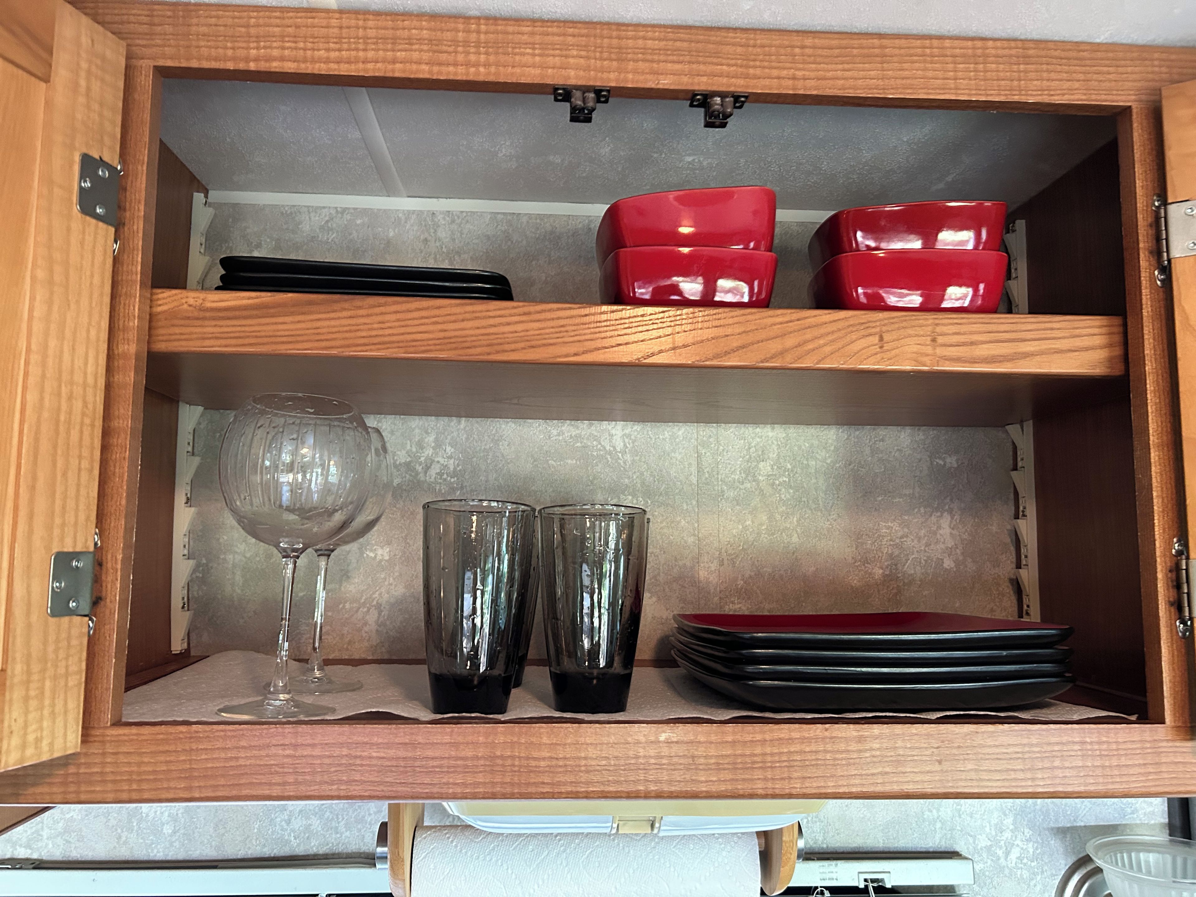 Un meuble en bois doté d’étagères contenant des récipients noirs et rouges, un verre à vin et deux verres transparents.