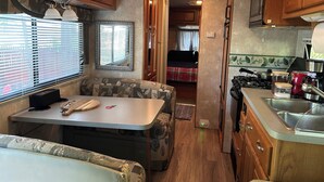 Cuina d'autocaravana compacta amb armaris de fusta, zona de menjador amb taula i banqueta, i fogó visible.