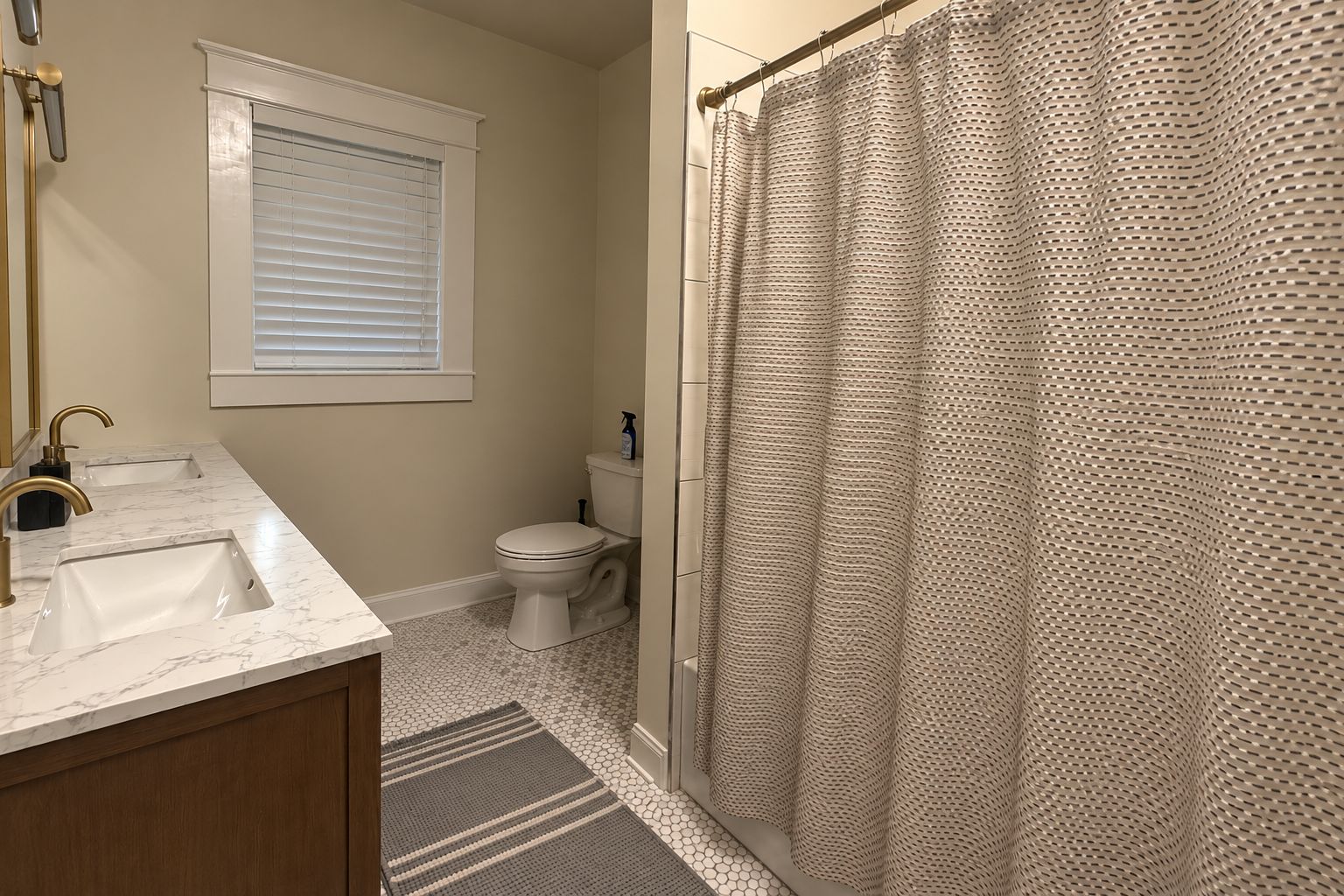 A bathroom with two sinks, a toilet, a shower curtain, and a window with blinds.