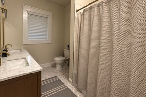 A bathroom with two sinks, a toilet, a shower curtain, and a window with blinds. - The Historic House (Dothan)