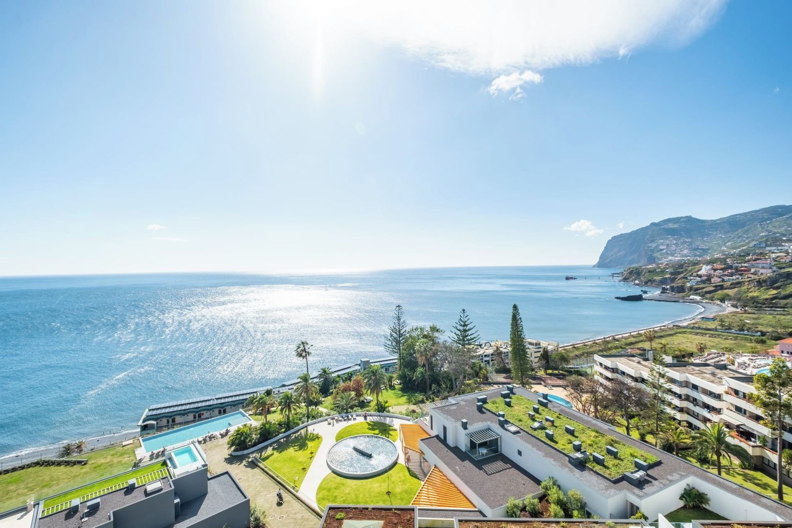 A coastal hotel with a swimming pool, a circular fountain, and a view of the sea.
