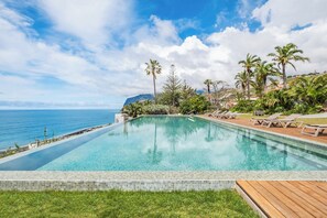 Ein rechteckiger Infinity-Pool mit Holzterrasse, umgeben von Palmen und Liegestühlen, mit Blick auf eine Küstenlandschaft.