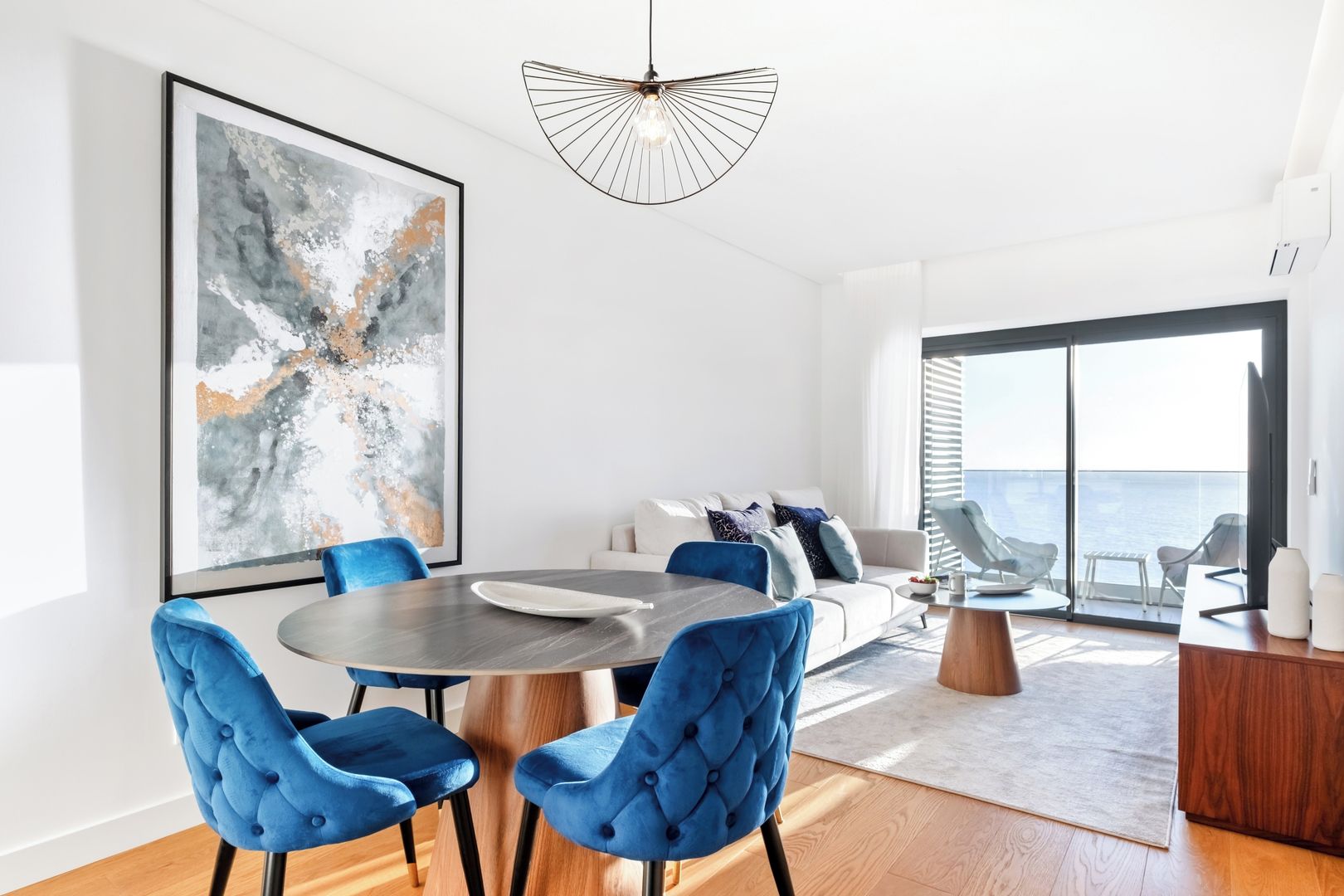 Un salon moderne avec une table à manger ronde, des chaises recouvertes d’un tissu bleu, un grand tableau abstrait, un canapé avec des coussins bleus et une vue sur la mer par de grandes fenêtres.