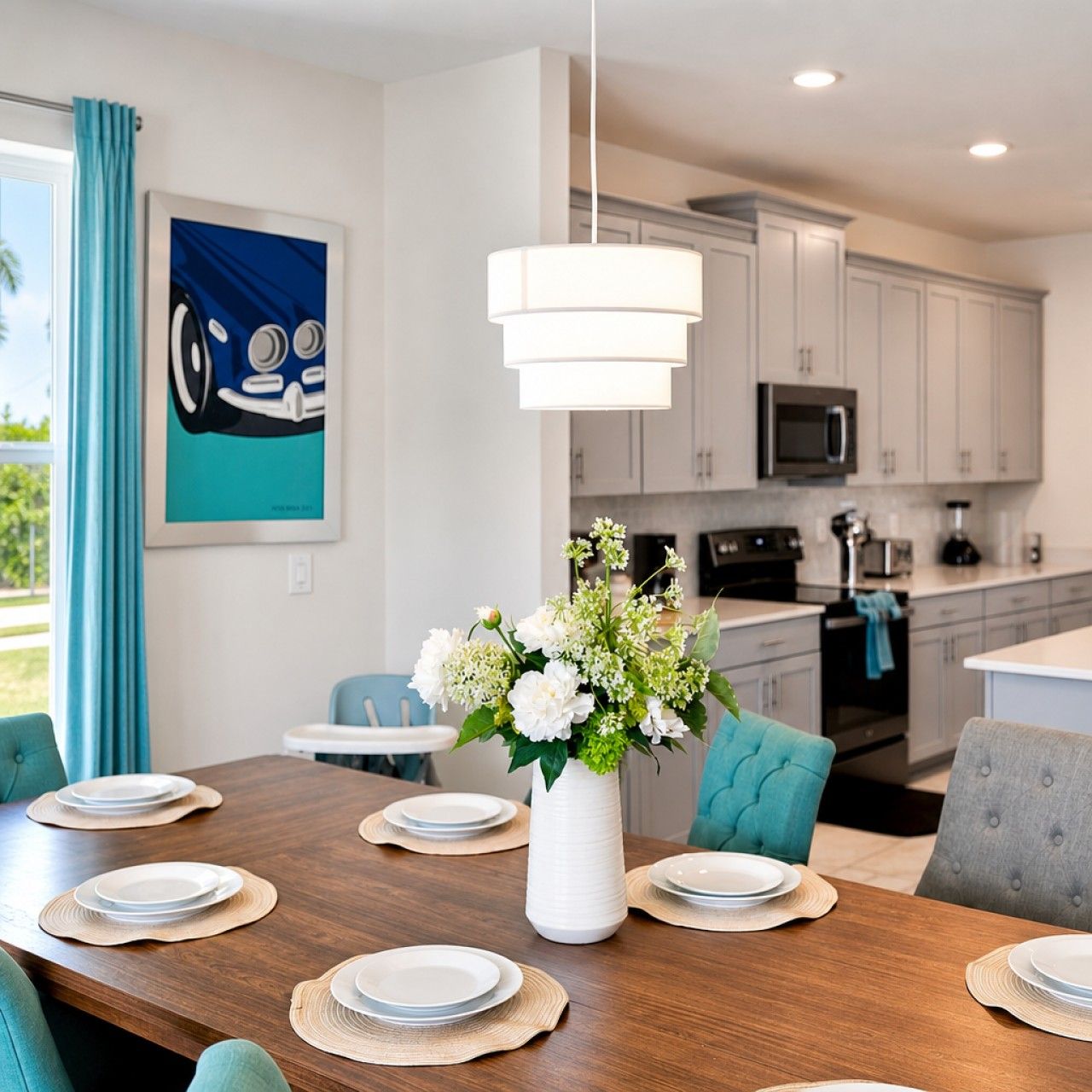 A dining area with a wooden table set for six, a white vase with flowers, and a kitchen in the background.