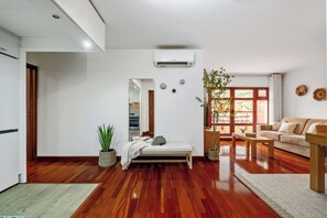 Ein modernes Wohnzimmer mit einer Couch, einem Couchtisch und einem Esstisch. Man sieht eine Pflanze, eine Bank und eine kleine Küchenzeile.