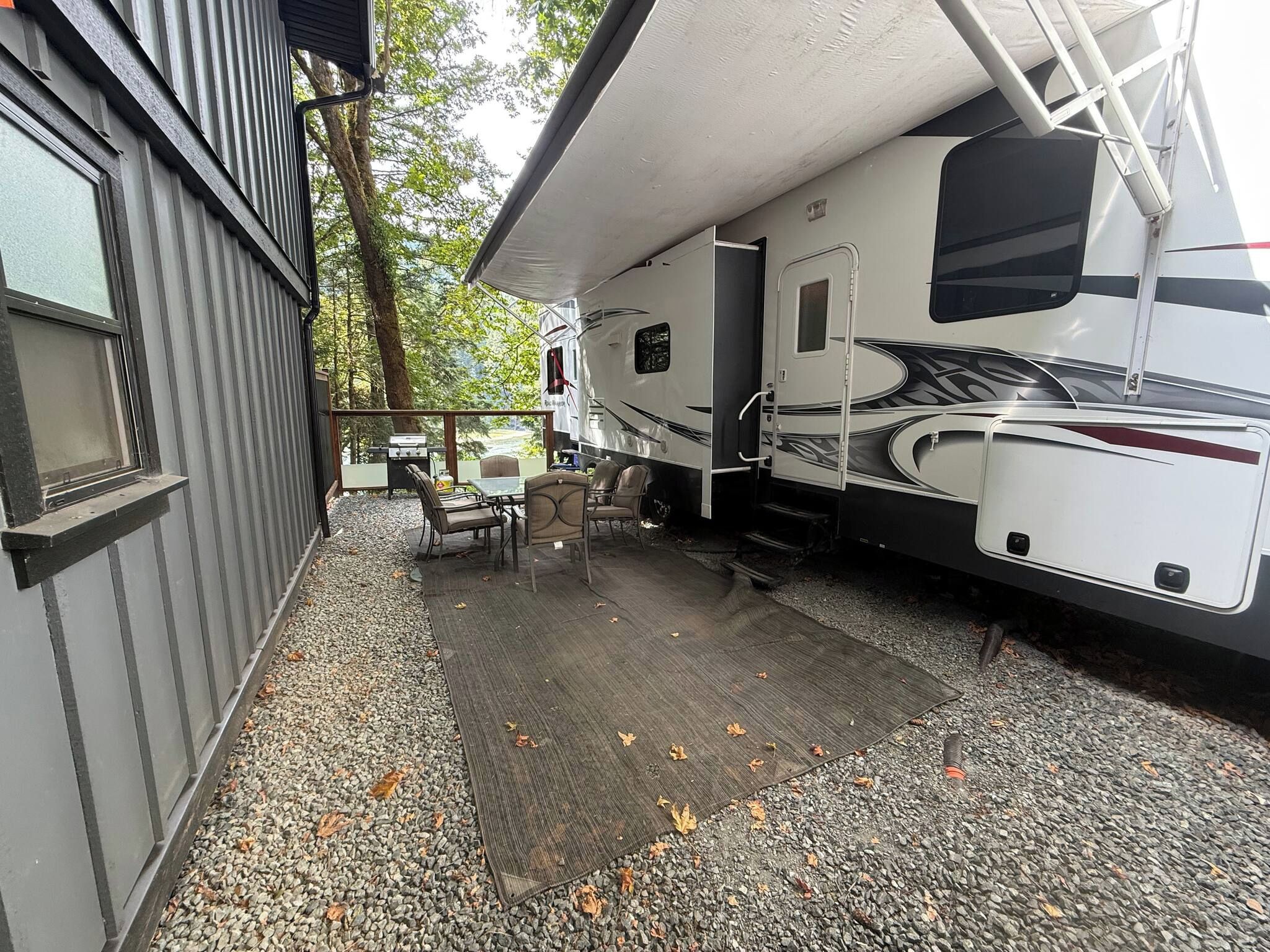 A covered outdoor area with a grill, seating, and a large RV parked adjacent to a building.