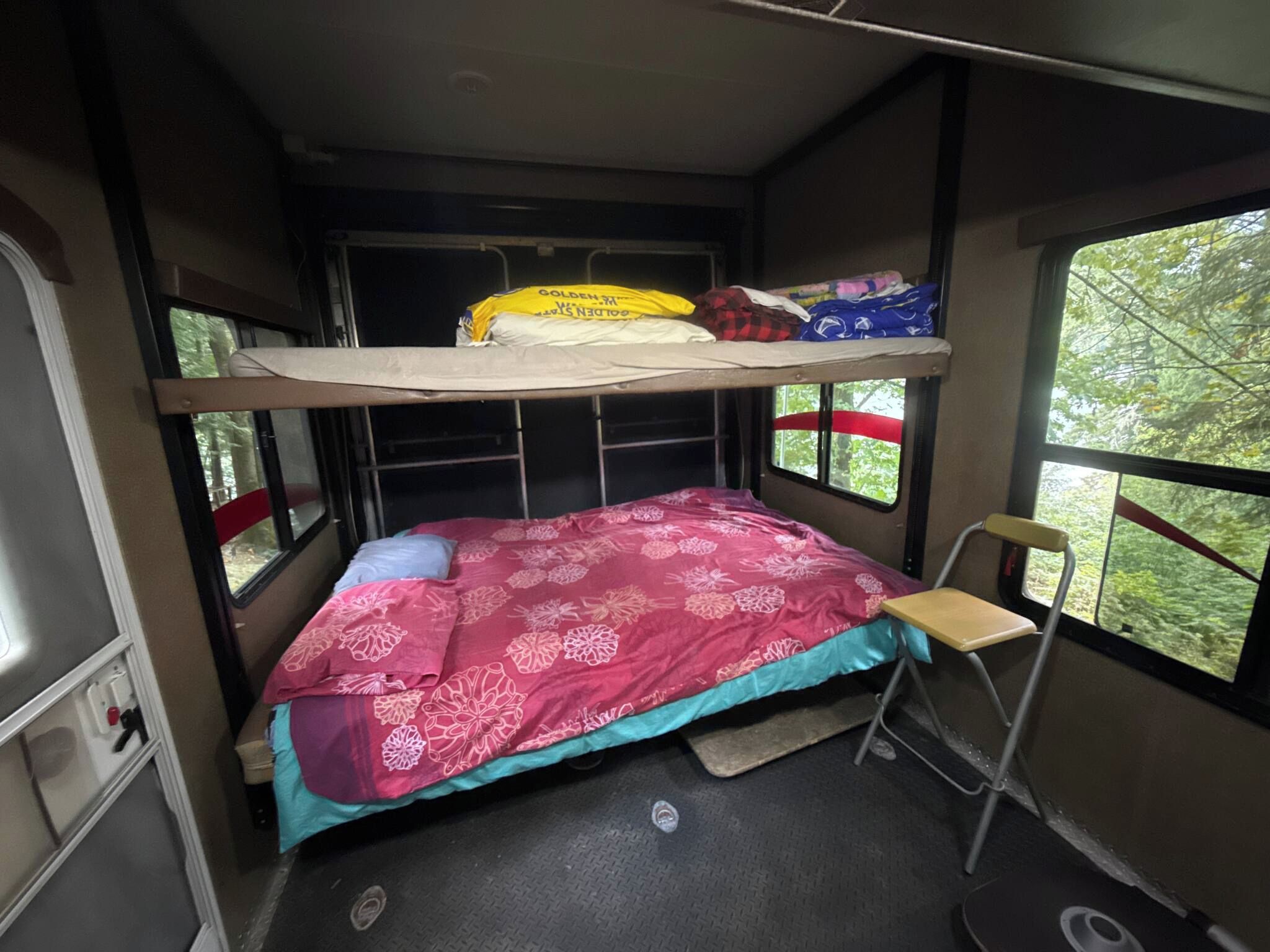 A compact, single-bunk RV interior with a bed, a small table, and a window providing a view of greenery outside.