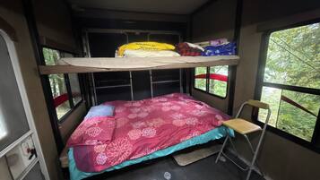 A compact, single-bunk RV interior with a bed, a small table, and a window providing a view of greenery outside.