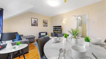 A modern living room with a dining area, grey sofas, a flat-screen TV, and a bookshelf.