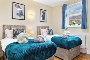 A hotel room with two beds, each with a blue blanket and a white towel. There are framed pictures on the wall, a window with blue curtains, and a radiator. - Huge 9BR London Stay Perfect For Groups, Near Leytonstone Tube & Stratford! (Greater London)