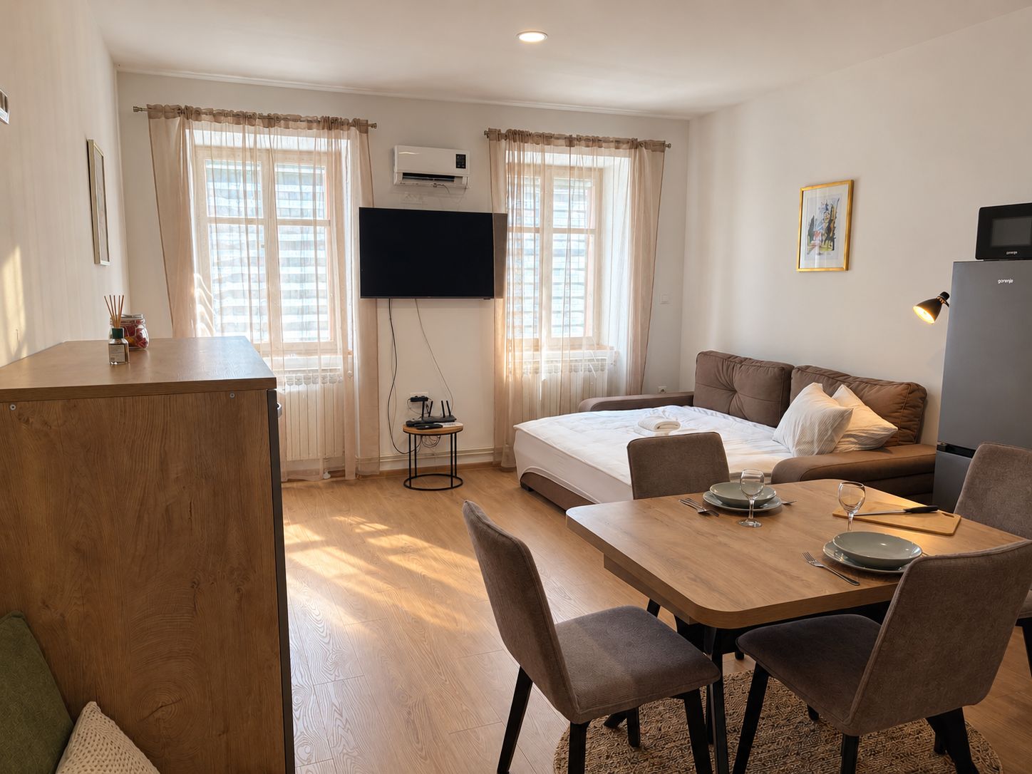 A hotel room with a bed, a sofa, a dining table with chairs, a TV, and an air conditioning unit.