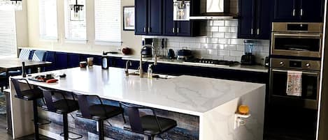 Una cocina moderna con armarios azul oscuro, una isla de mármol blanco con taburetes de bar, electrodomésticos de acero inoxidable y un amplio ventanal con persianas.