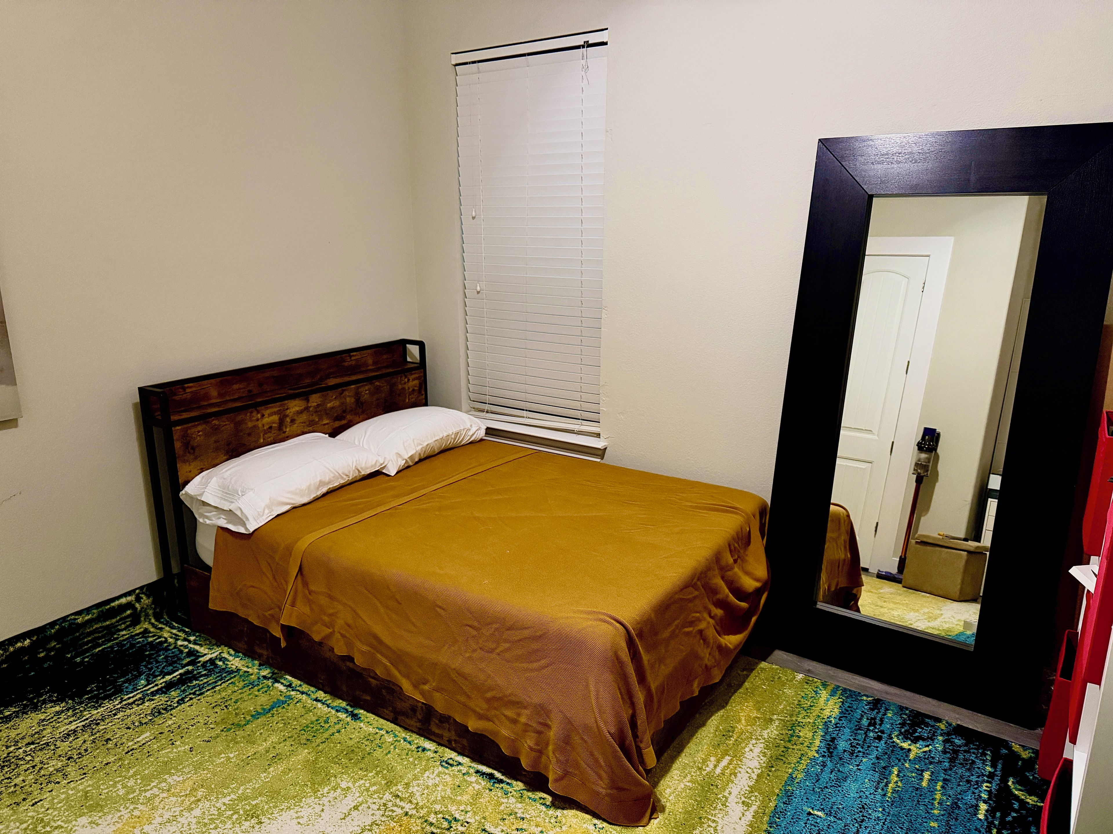 A single bed with a wooden headboard, a white pillow, and a mustard-colored bedspread. A window with blinds is visible to the left of the bed.