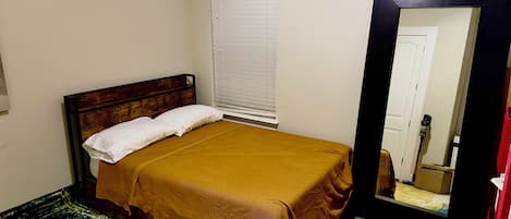 A single bed with a wooden headboard, a white pillow, and a mustard-colored bedspread. A window with blinds is visible to the left of the bed.