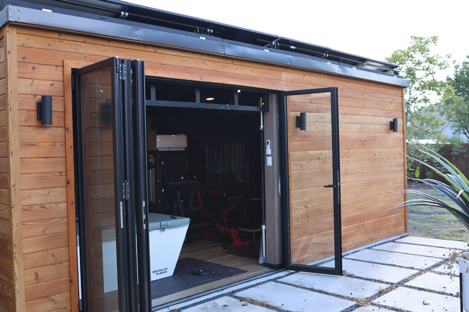 A wooden structure with a glass door, a visible interior with a mattress labeled 'Brooklyn Plunge,' and a metal roof.