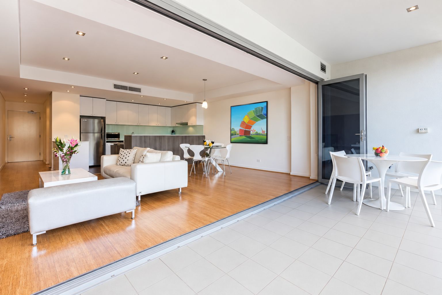 A modern living room with a sofa, dining table, and chairs. There is a kitchen area with cabinets and a refrigerator.