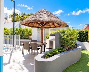 Eine Terrasse im Freien mit einem Reetdachschirm, einem Grill und einer Sitzgruppe mit Stühlen.