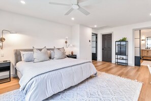 Una camera da letto con un letto, comodini, un tavolo di fianco al letto e una ventola a soffitto, con una porta che dà su un'altra stanza.