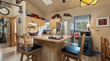A kitchen with wooden cabinets, a stainless steel refrigerator, a wooden island with chairs, and a hanging light fixture.