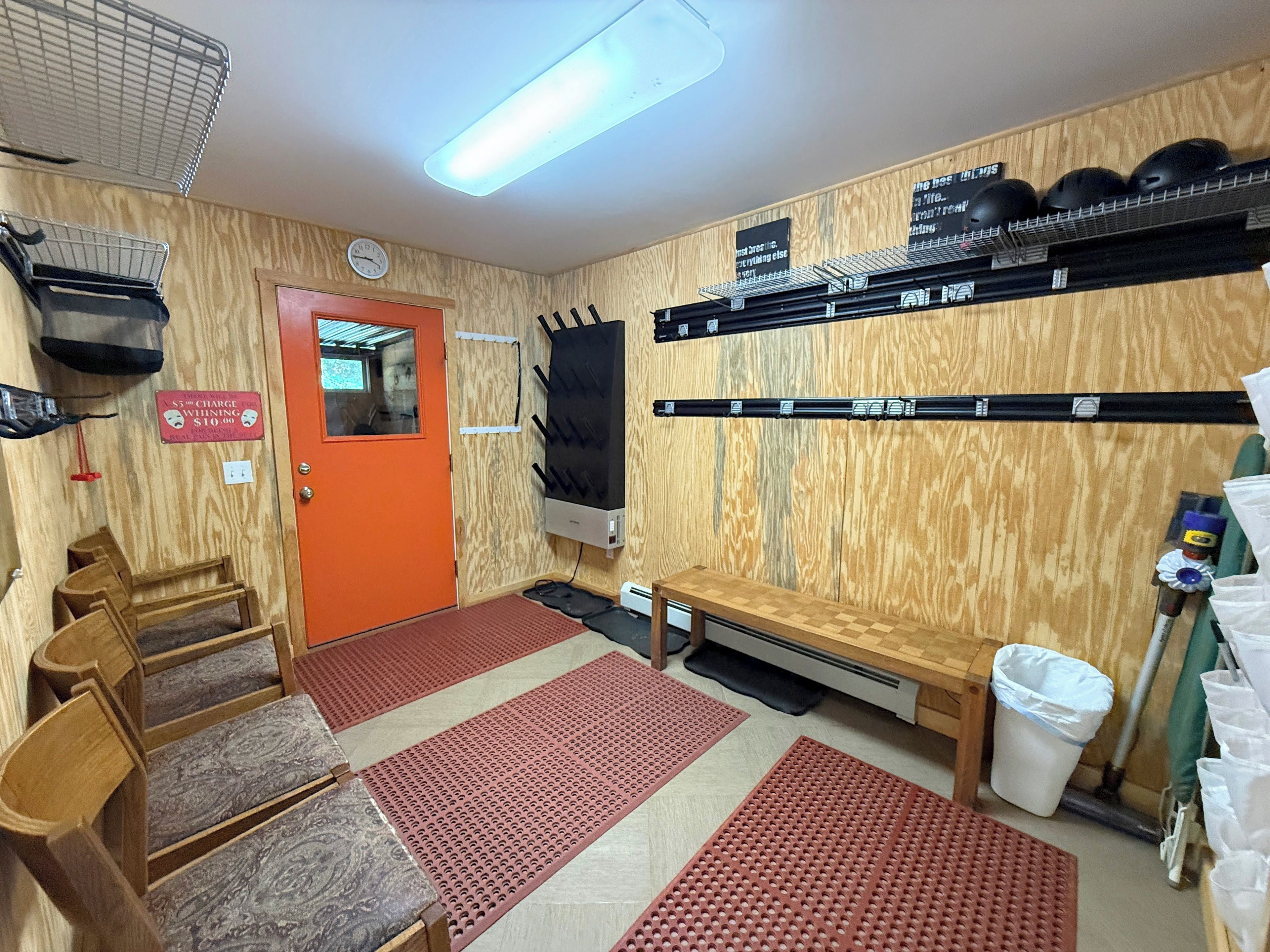 A room with a wooden wall, a red door, a bench, a trash bin, and shelves with helmets.