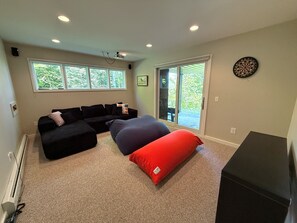 Un salon avec un canapé d’angle, un coussin rouge, un téléviseur à écran plat et un jeu de fléchettes accroché au mur.