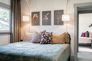 En snyggt bäddad säng med ett blått överkast och flera kuddar, ett fönster med gardiner och tre inramade blomstermotiv på väggen.