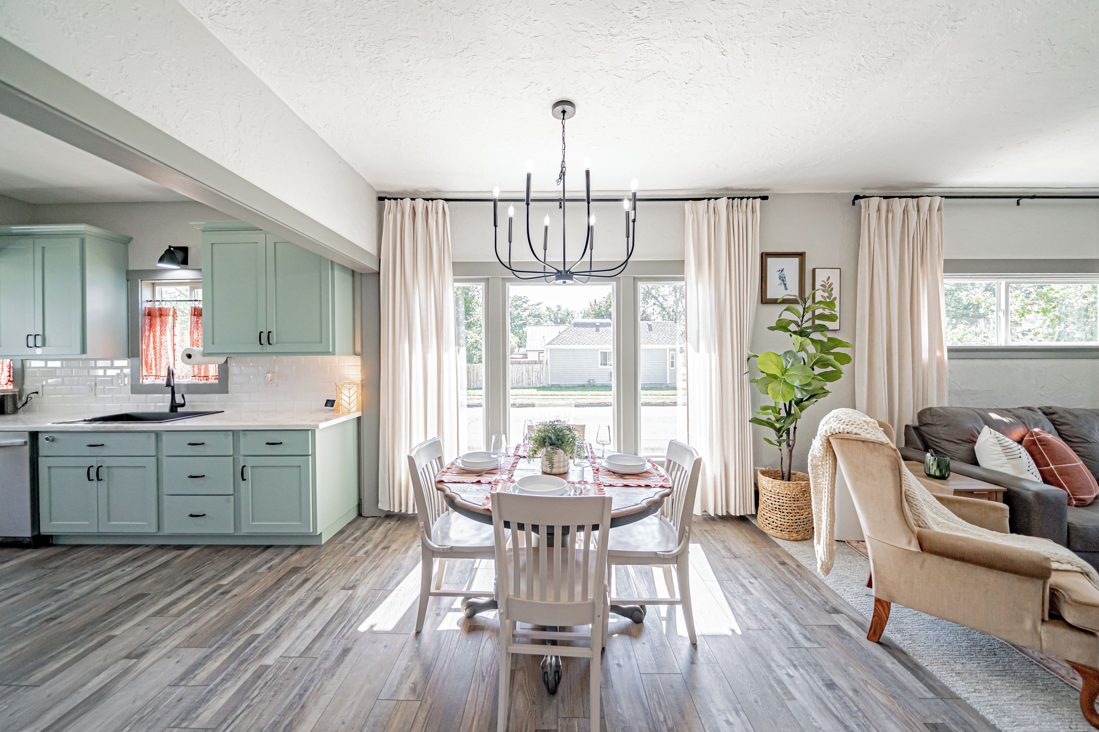 A spacious kitchen with light green cabinets, a dining area with a table set for four, and a living area with a sofa and a plant.