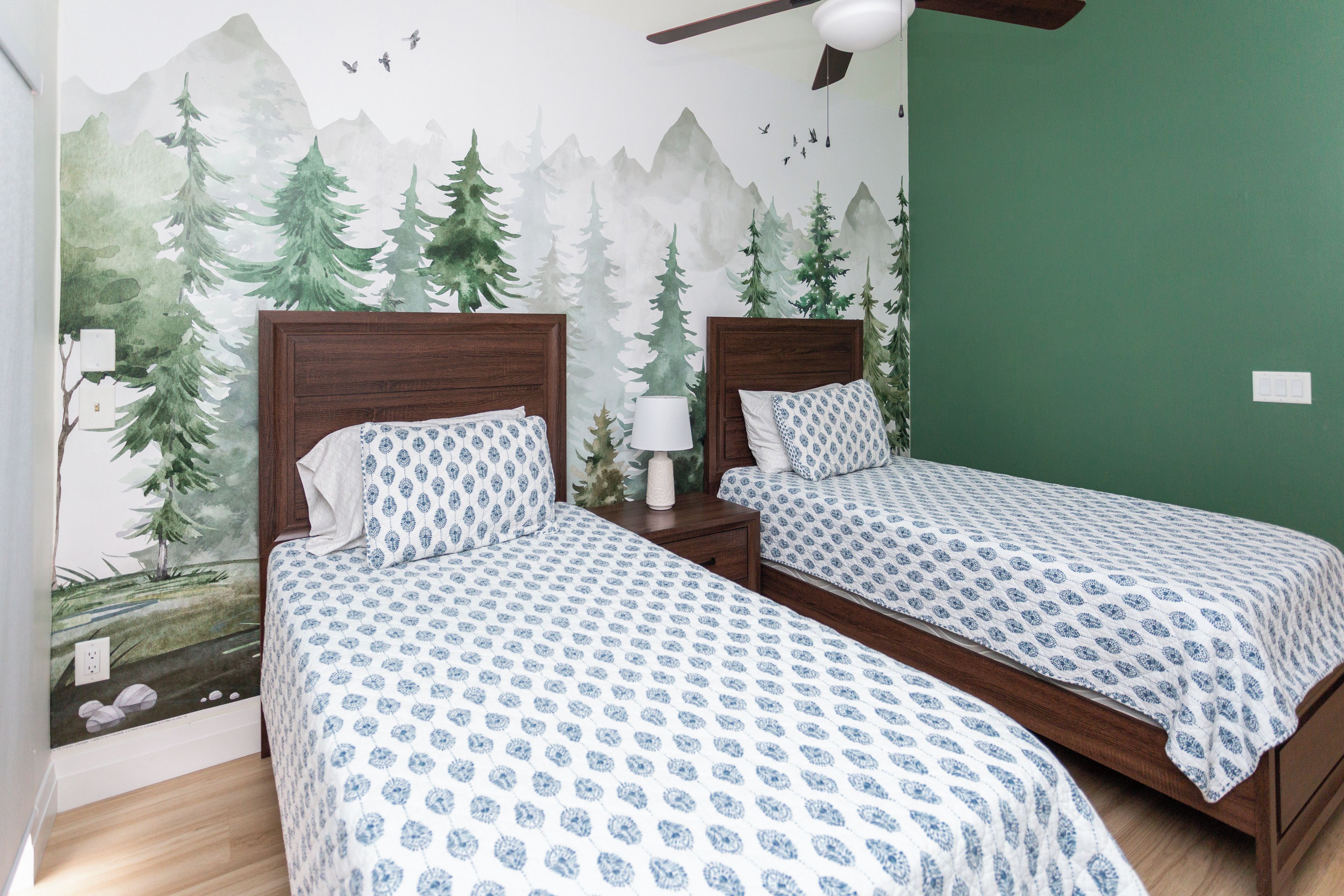 A room with two beds, each with a patterned bedspread, a nightstand with a lamp, and a wall mural of a forest scene.