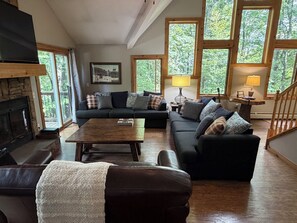 A living room with a fireplace, a sofa set, a coffee table, and large windows with a view of trees.
