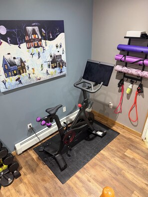 A room with a stationary bike, a wall-mounted TV, and a painting of a ski lodge.