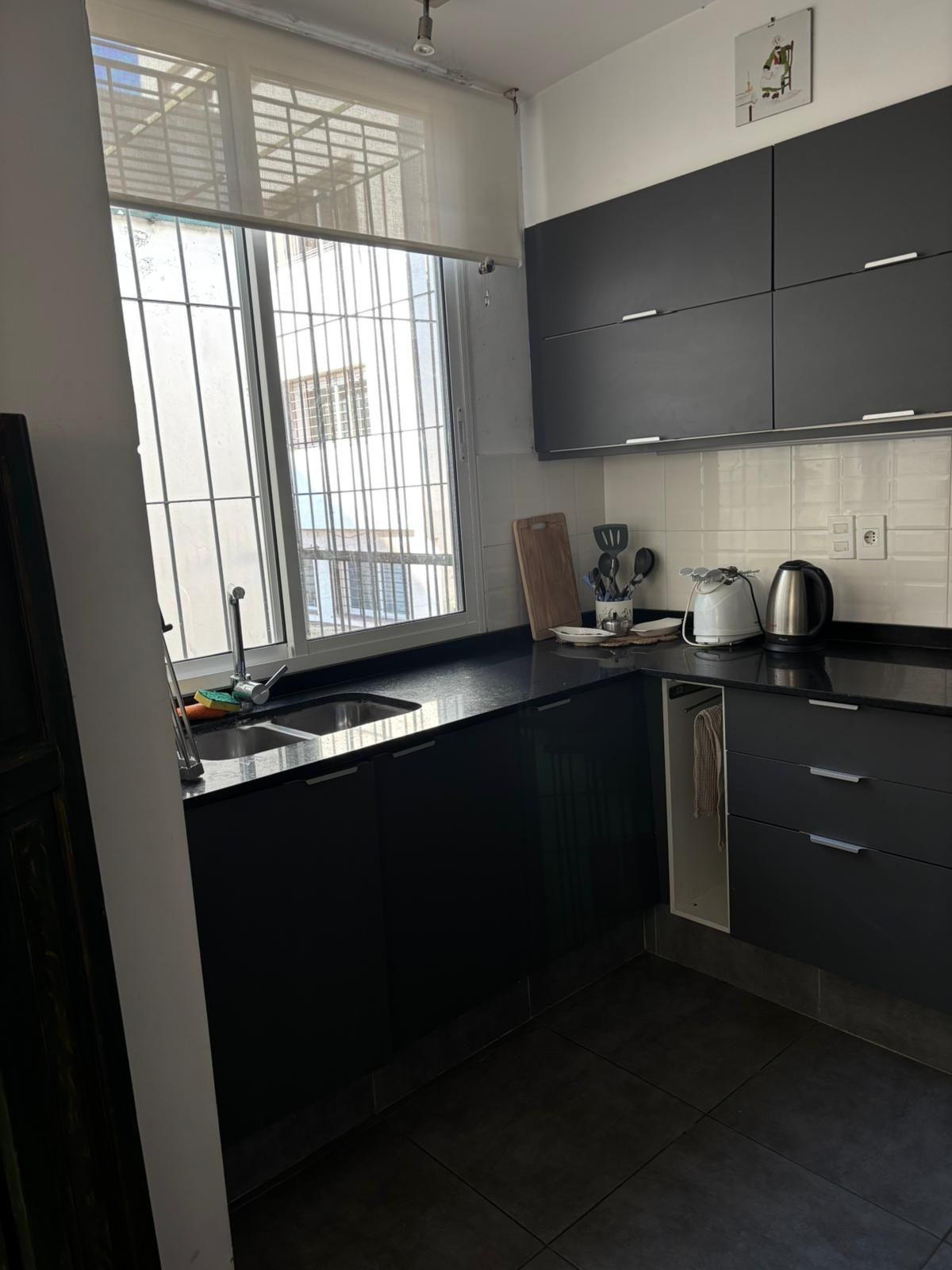 A modern kitchen with dark cabinetry, a stainless steel sink, and a window with a view of buildings outside.