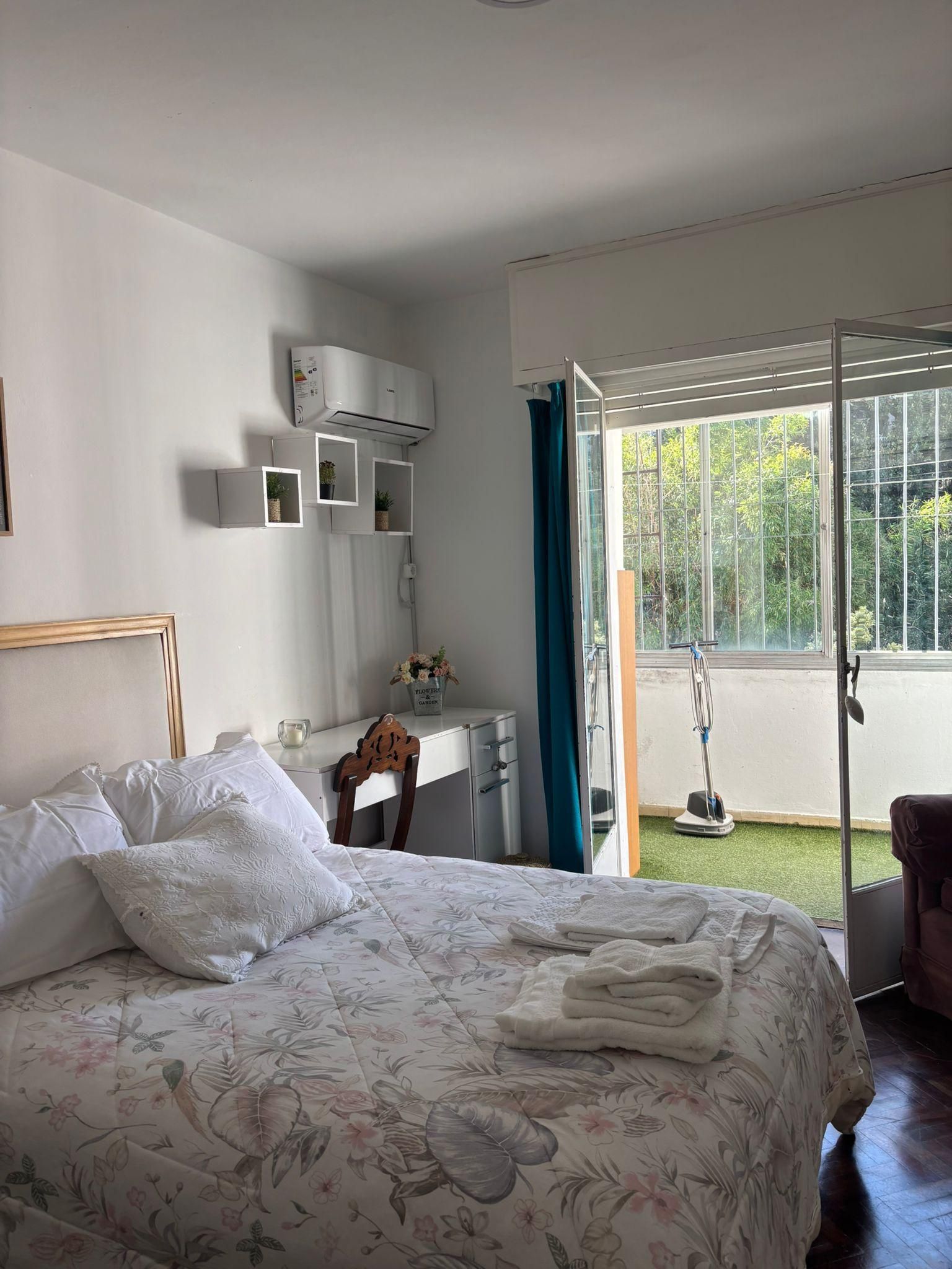 A bedroom with a bed, a desk, a window with a view of greenery, and an air conditioning unit.