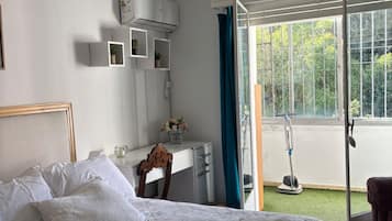 A bedroom with a bed, a desk, a window with a view of greenery, and an air conditioning unit.
