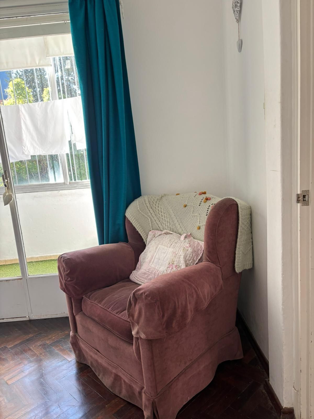 A corner room with a pink armchair, a white knitted throw, and a teal curtain.