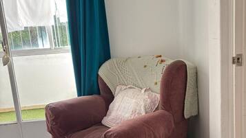 A corner room with a pink armchair, a white knitted throw, and a teal curtain.