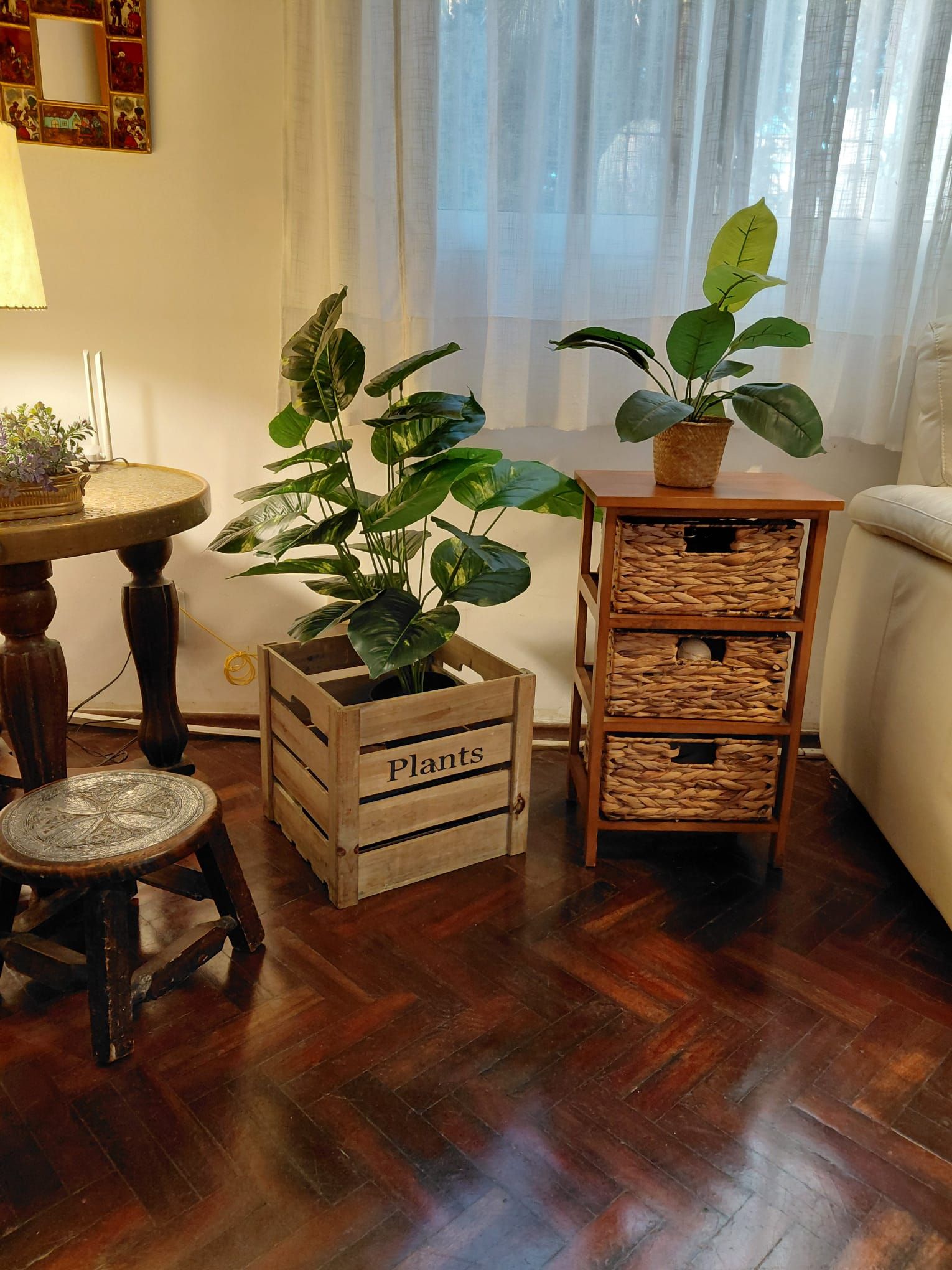 A room with a wooden floor, a round table with a lamp, a wooden stool, and two wooden crates labeled 'Plants' containing potted plants.