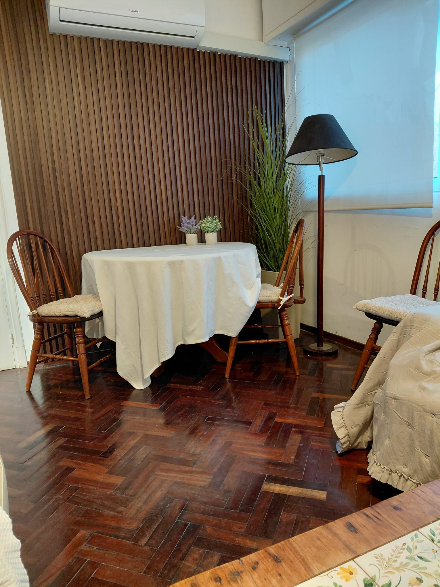 A dining area with a wooden herringbone floor, a round table with a white tablecloth, two wooden chairs, a potted plant, and a floor lamp.