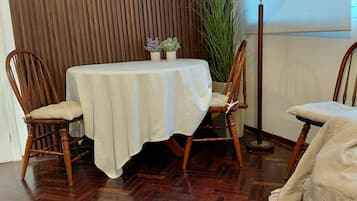 A dining area with a wooden herringbone floor, a round table with a white tablecloth, two wooden chairs, a potted plant, and a floor lamp.