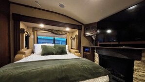 A bedroom with a bed, a television, a fireplace, and a window with blinds. - Well stocked 5th wheel and private space at Christina Lake Resort. (Christina Lake)
