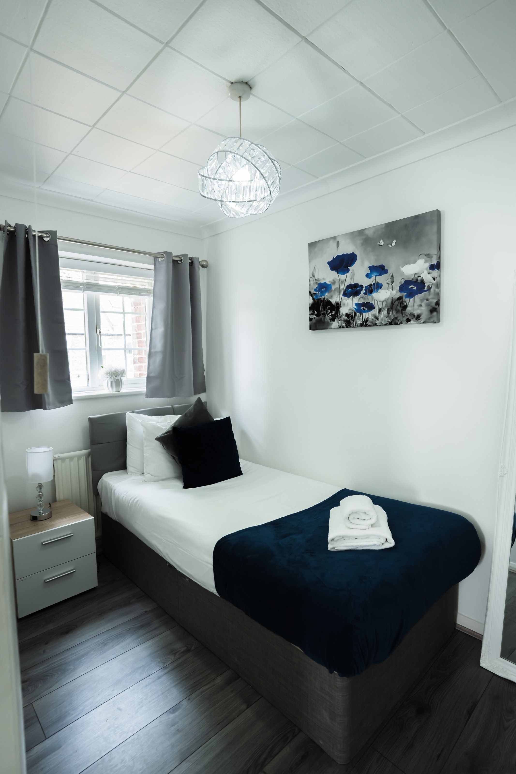 Une chambre à coucher avec un lit, une table de chevet, une lampe, une fenêtre avec des rideaux et un tableau au mur.
