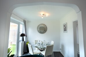 A dining area with a glass table, white chairs, a decorative mirror, and a view of the outdoors. - 3-Bed Hse in Central Windsor w/Garden (Windsor)