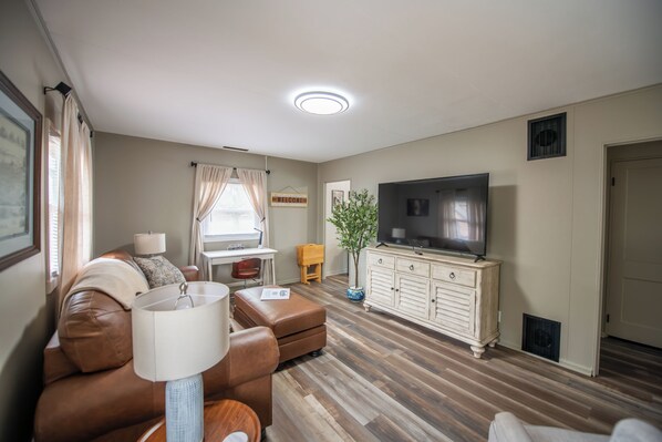 Una sala de estar con un sofá de cuero marrón, un televisor sobre un mueble blanco y piso de madera.