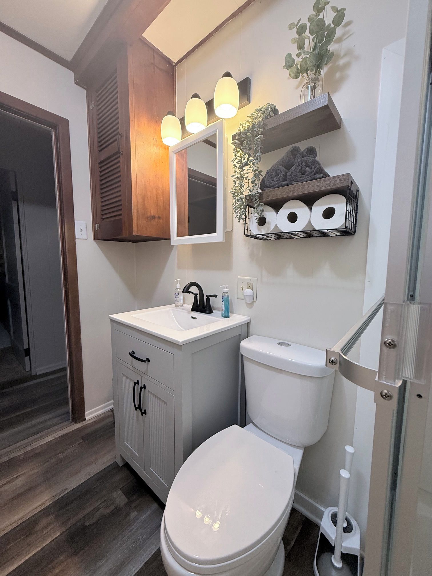 Een badkamer met een wit wastafelmeubel, een toilet en een spiegel boven het wastafelmeubel. Boven de spiegel hangen drie lampen en er is een plank met handdoeken en toiletpapier.
