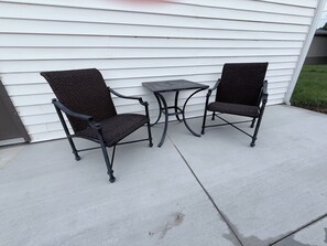 Dos sillas de mimbre y una pequeña mesa cuadrada en un patio de concreto.