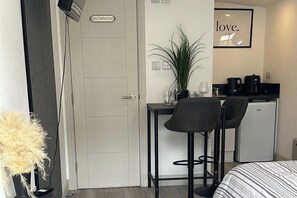 A hotel room with a bed, a desk with a chair, a small refrigerator, a potted plant, and a sign indicating a bathroom. - Studio Sleeps 2 with WiFi (Thornton Heath)