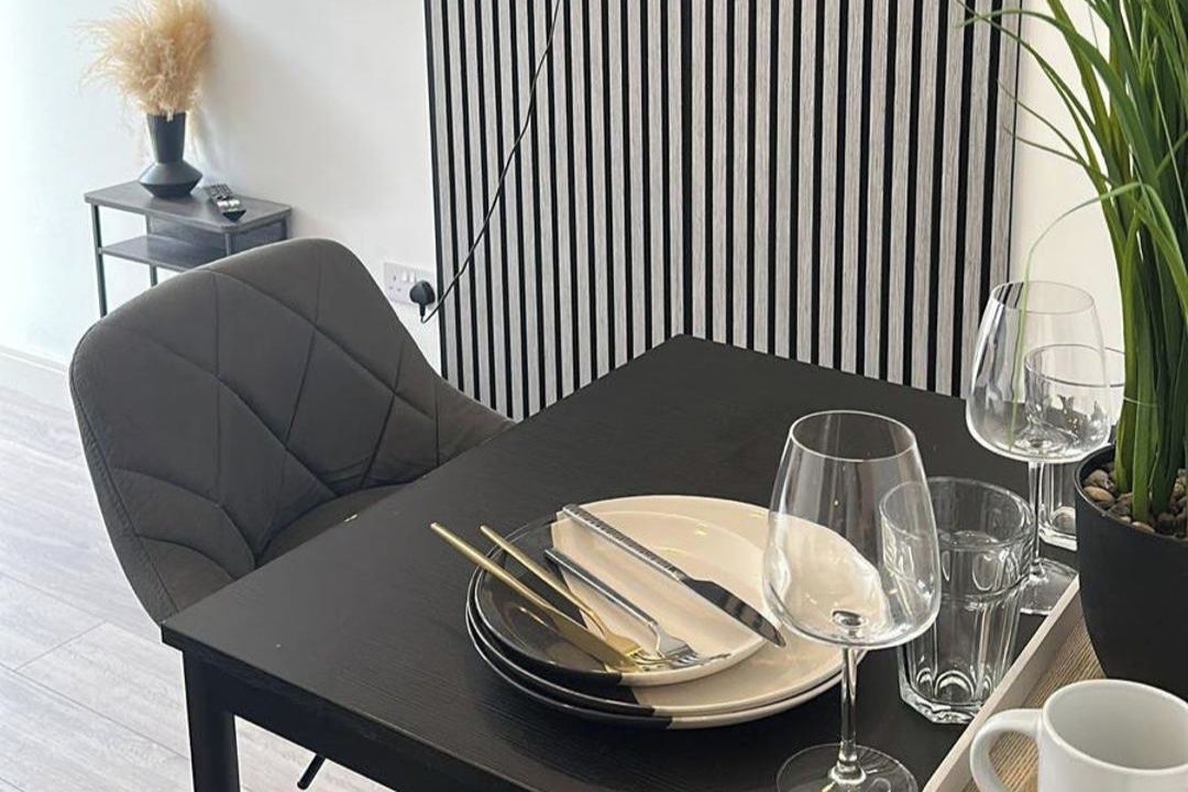 Et spisebord dækket med tallerkener, bestik og glas, sammen med en stol og et lille sofabord med en vase og en fjernbetjening.