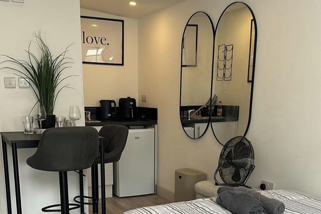 Une chambre d’hôtel compacte comprenant un lit, un bureau avec deux chaises, une petite kitchenette et un grand miroir.