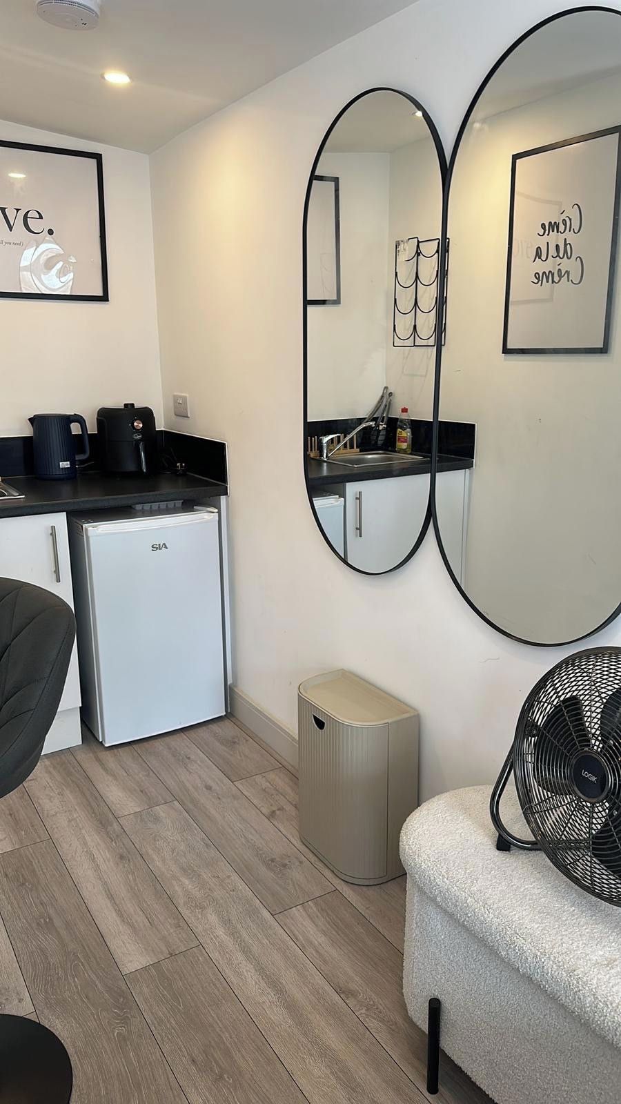 Una kitchenette compacta con refrigerador, cafetera y hervidor. Hay dos espejos en la pared, un basurero pequeño y un ventilador.